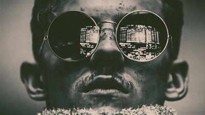 A close-up portrait of a man wearing round sunglasses, reflecting a vibrant cityscape in the lenses.