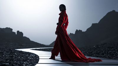 A woman in a flowing red dress walks along a winding path in a desolate, rocky landscape.