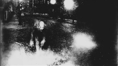 A grainy, black and white photograph depicts a figure seemingly running towards the viewer, set against a blurred, nighttime backdrop.