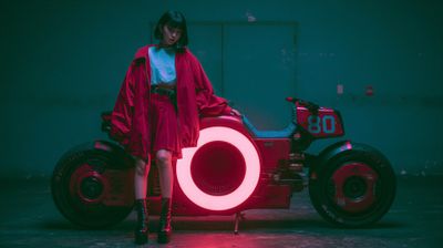 A stylish woman poses confidently beside a futuristic motorcycle in a dimly lit, industrial setting.