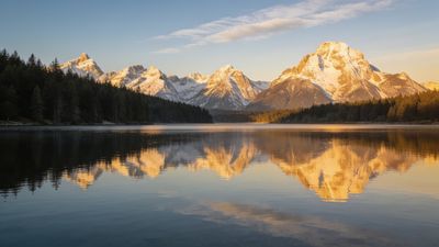 A tranquil scene of snow-capped mountains reflecting on a calm lake at sunset, with the warm glow of the setting sun casting a golden hue over the peaks and surrounding forest, creating a serene and peaceful atmosphere.