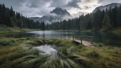 A tranquil mountain lake is surrounded by dense evergreen forests, with a wooden bridge crossing over a small inlet, set against the backdrop of mist-covered peaks under a cloudy sky, creating a serene and peaceful atmosphere.