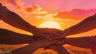A group of hands gently stack together against a vibrant sunset backdrop, symbolizing unity and friendship. The warm hues of orange and pink in the sky create a serene and hopeful atmosphere.
