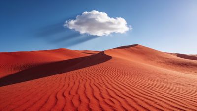 A vast expanse of red desert dunes stretches under a clear blue sky, with a single fluffy white cloud casting a soft shadow, creating a tranquil and serene atmosphere.