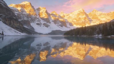A golden sunrise casts a warm glow over snow-covered mountain peaks, reflecting in a tranquil lake below, creating a serene and majestic atmosphere.