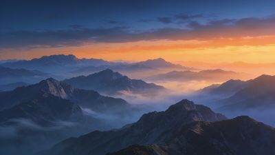 A breathtaking sunrise casts a warm golden glow over a series of mist-covered mountain peaks, creating a serene and majestic atmosphere as the sky transitions from deep blue to vibrant orange.
