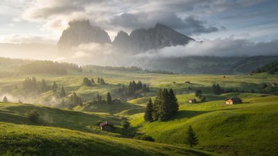 A serene mountain landscape features towering peaks shrouded in mist, with lush, rolling green hills dotted with small wooden cabins in the foreground, creating a tranquil and picturesque scene.