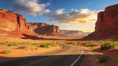 A winding road stretches through a vast desert landscape, flanked by towering red rock formations under a vibrant sunset sky, creating a serene and majestic atmosphere.