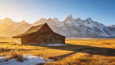 A rustic wooden barn stands in a sunlit field with a backdrop of snow-capped mountains under a clear blue sky, creating a serene and picturesque landscape.