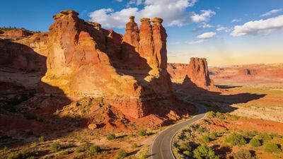 A winding road cuts through a vast desert landscape, flanked by towering red rock canyons under a clear blue sky, creating a serene and majestic atmosphere.