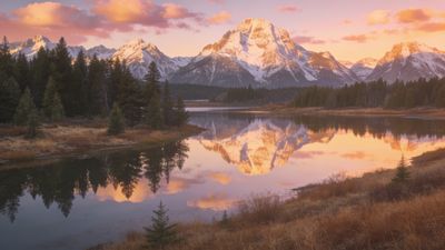 A tranquil scene of snow-capped mountains is reflected in a calm lake at sunset, surrounded by dense forests and golden grasslands, creating a peaceful and serene atmosphere.