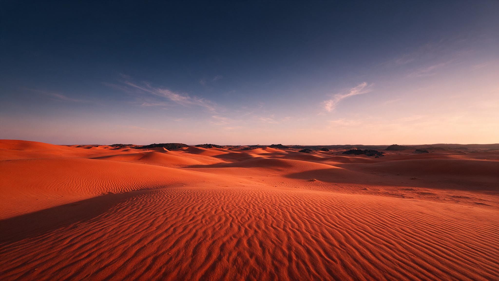 Rolling red-orange dunes of the Sahara Desert stretch to the horizon beneath a cloud-streaked sky lit by the setting sun. The vast, empty landscape and soft evening light create a quiet, tranquil mood.