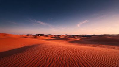 Rolling red-orange dunes of the Sahara Desert stretch to the horizon beneath a cloud-streaked sky lit by the setting sun. The vast, empty landscape and soft evening light create a quiet, tranquil mood.