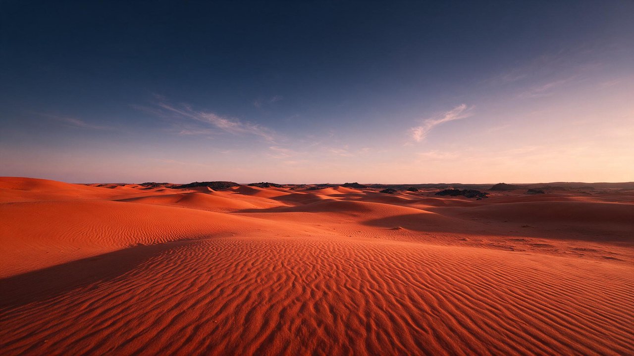 Rolling red-orange dunes of the Sahara Desert stretch to the horizon beneath a cloud-streaked sky lit by the setting sun. The vast, empty landscape and soft evening light create a quiet, tranquil mood.