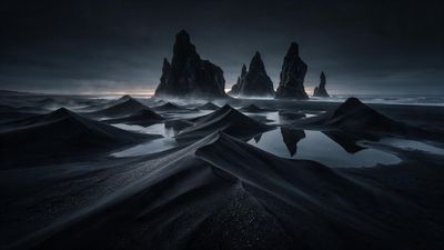 Jagged Reynisdrangar basalt sea stacks rise from the ocean beyond rippling black sand dunes and reflective tide pools on Iceland’s south coast at dusk, shrouded in mist. The low, dim light and dark tones create a brooding, tranquil mood.