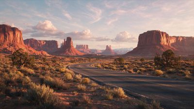 A winding asphalt road curves into Monument Valley, flanked by scrub desert and towering red sandstone buttes. The warm sunset glow and pastel clouds evoke a calm, open-road serenity.