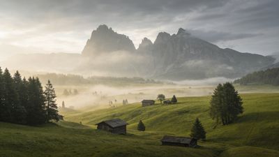 A serene morning scene in the Alps features mist rolling over lush green hills, dotted with rustic wooden cabins, while towering mountain peaks rise majestically in the background under a soft, cloudy sky. The mood is tranquil and picturesque, evoking a sense of peaceful solitude.