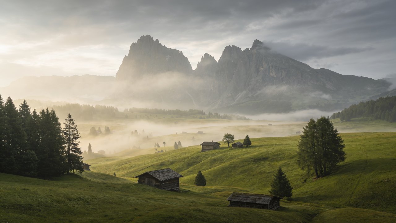A serene morning scene in the Alps features mist rolling over lush green hills, dotted with rustic wooden cabins, while towering mountain peaks rise majestically in the background under a soft, cloudy sky. The mood is tranquil and picturesque, evoking a sense of peaceful solitude.