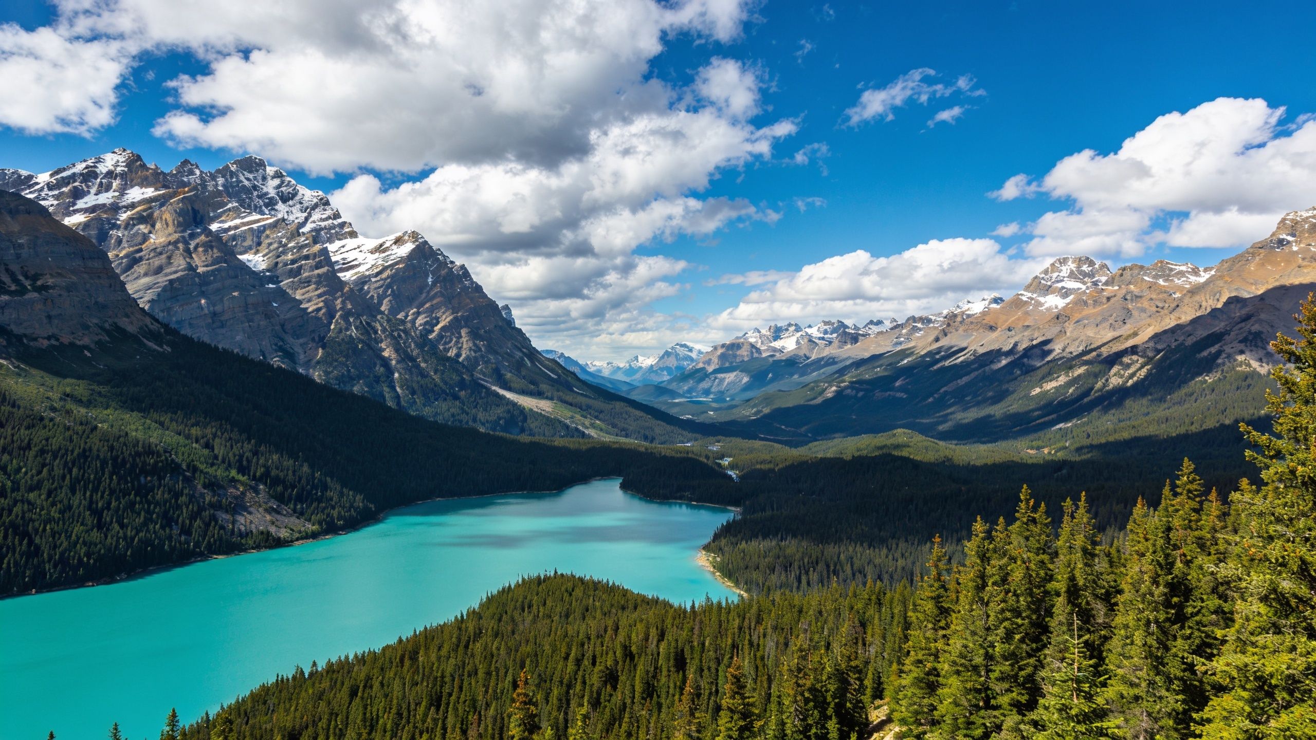 A serene turquoise lake is nestled among lush evergreen forests and towering snow-capped mountains under a clear blue sky, creating a tranquil and majestic atmosphere.