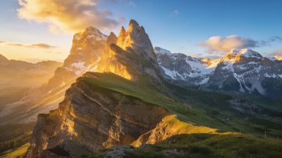 A golden sunrise bathes the jagged peaks of a majestic mountain range in warm light, casting dramatic shadows over lush green valleys and creating a serene, awe-inspiring atmosphere.