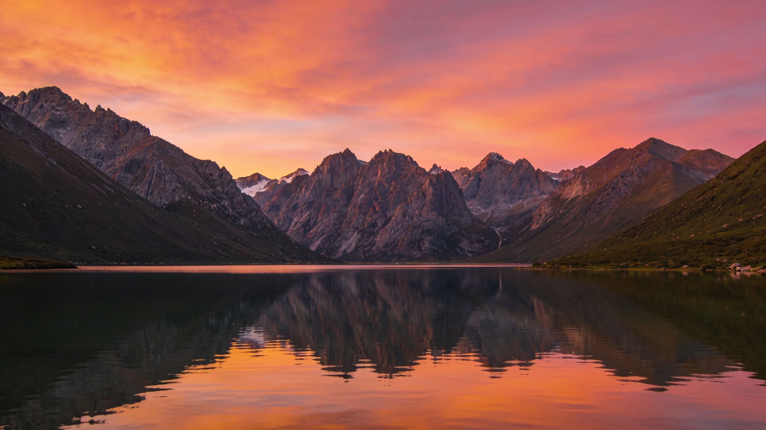 A serene mountain landscape is captured at sunset, with rugged peaks reflecting in a tranquil lake under a vibrant sky of orange and pink hues, creating a peaceful and majestic atmosphere.