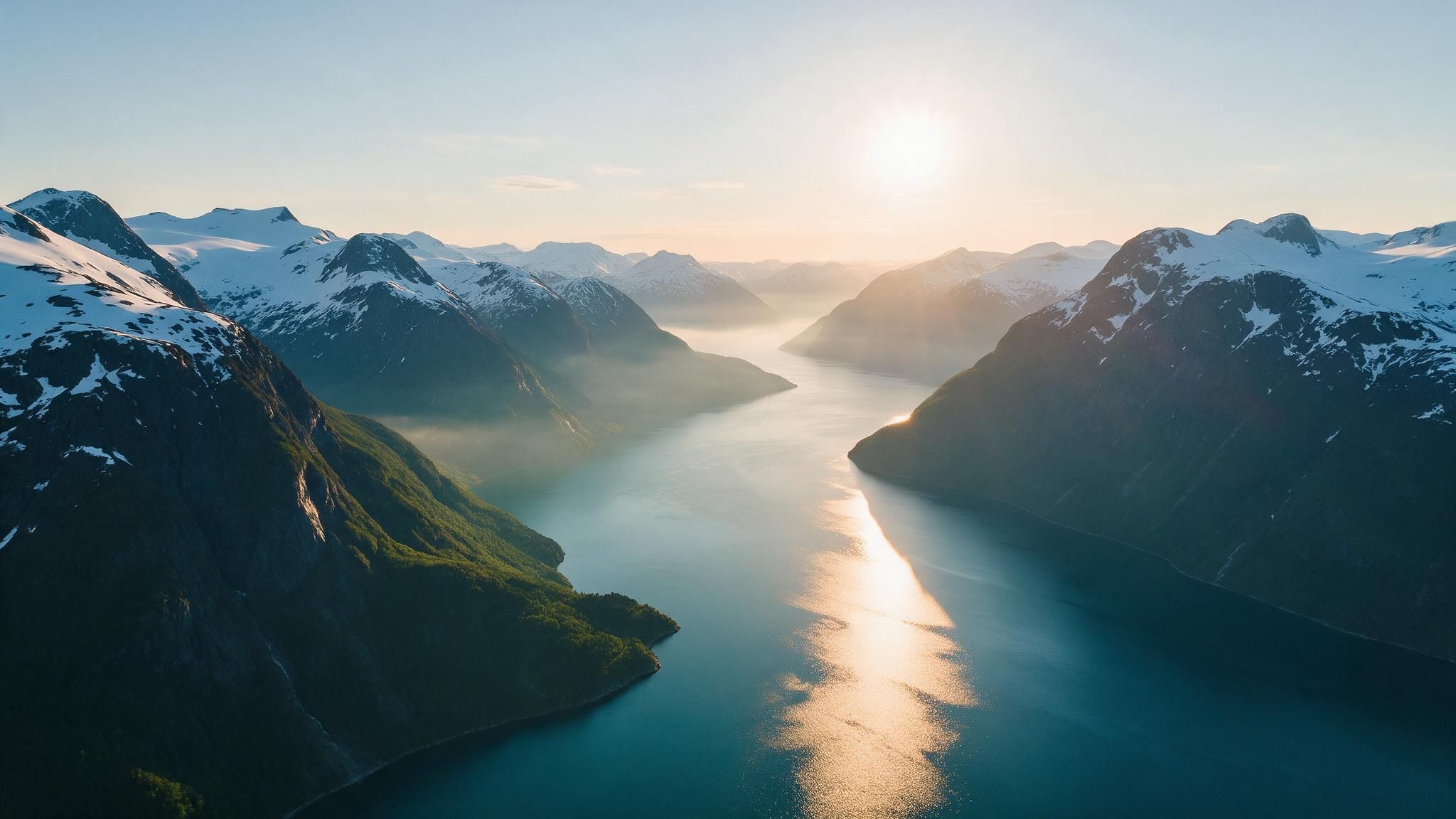 Aerial view of a Norwegian fjord framed by snow-capped mountains as the rising sun paints a golden path on the calm blue water, creating a serene, crisp morning mood.