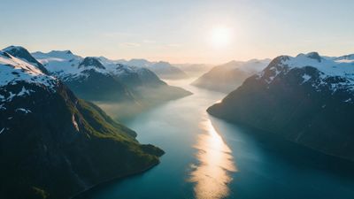 Aerial view of a Norwegian fjord framed by snow-capped mountains as the rising sun paints a golden path on the calm blue water, creating a serene, crisp morning mood.