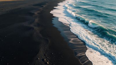Aerial view of Iceland’s black sand coastline where teal waves curl onto the dark shore, creating a stark contrast in a quiet, rugged seascape.